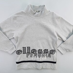 May include: A light gray sweatshirt with a mock neck collar. The word "ellesse PERUGIA" is printed across the chest in black and white. A black stripe is at the bottom of the sweatshirt.