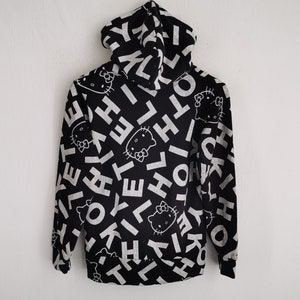 May include: Black hoodie with a white all-over print featuring the text "HELLO" and the Hello Kitty character. The hoodie has a full zipper and a hood. The design is repeated across the entire garment.