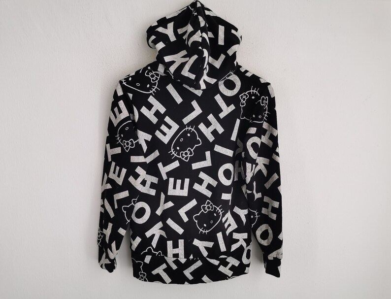 May include: Black hoodie with a white all-over print featuring the text "HELLO" and the Hello Kitty character. The hoodie has a full zipper and a hood. The design is repeated across the entire garment.