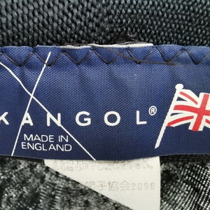 May include: A close-up of a navy blue label with the brand name "KANGOL" in white letters. The label also features the text "MADE IN ENGLAND" and a Union Jack flag. The label is stitched onto a black fabric.