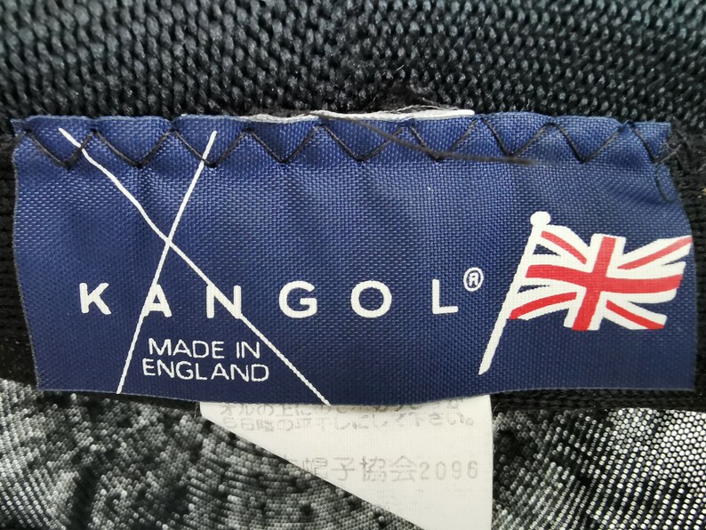 May include: A close-up of a navy blue label with the brand name "KANGOL" in white letters. The label also features the text "MADE IN ENGLAND" and a Union Jack flag. The label is stitched onto a black fabric.