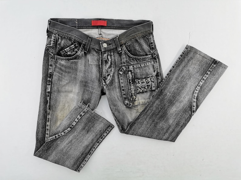 May include: Faded black denim jeans with a distressed look. Features include multiple pockets, a button and zipper closure, and a red tag on the waistband. The jeans are laid flat on a white surface.