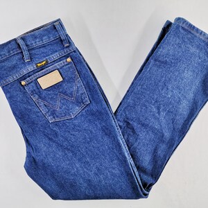 Puede incluir: Vaqueros de mezclilla azul oscuro con un dise&ntilde;o cl&aacute;sico de cinco bolsillos. Cuenta con una etiqueta Wrangler en el bolsillo trasero y un parche de cuero. Los vaqueros est&aacute;n doblados cuidadosamente, mostrando los detalles de las costuras y la textura de la mezclilla.