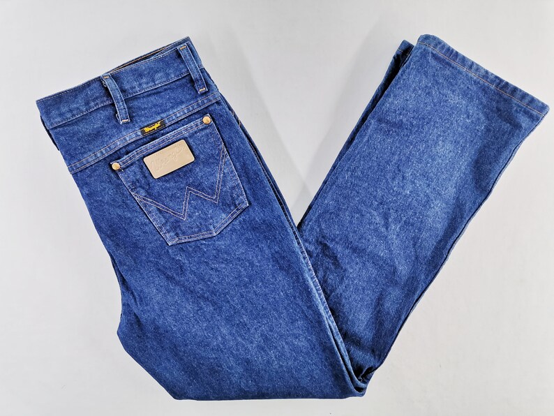 Puede incluir: Vaqueros de mezclilla azul oscuro con un dise&ntilde;o cl&aacute;sico de cinco bolsillos. Cuenta con una etiqueta Wrangler en el bolsillo trasero y un parche de cuero. Los vaqueros est&aacute;n doblados cuidadosamente, mostrando los detalles de las costuras y la textura de la mezclilla.