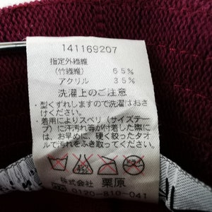 K&ouml;nnte beinhalten: Nahaufnahme eines weinroten Stoffes mit einem wei&szlig;en Etikett. Das Etikett enth&auml;lt japanischen Text, einschlie&szlig;lich Prozents&auml;tzen f&uuml;r Bambusfaser und Acryl. Waschanweisungen und ein Pflegeetikett sind ebenfalls sichtbar.