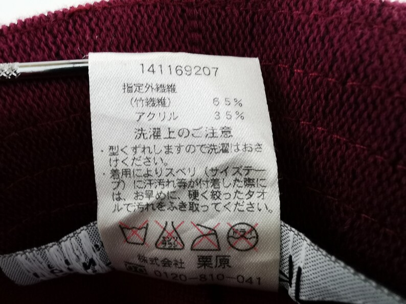 K&ouml;nnte beinhalten: Nahaufnahme eines weinroten Stoffes mit einem wei&szlig;en Etikett. Das Etikett enth&auml;lt japanischen Text, einschlie&szlig;lich Prozents&auml;tzen f&uuml;r Bambusfaser und Acryl. Waschanweisungen und ein Pflegeetikett sind ebenfalls sichtbar.