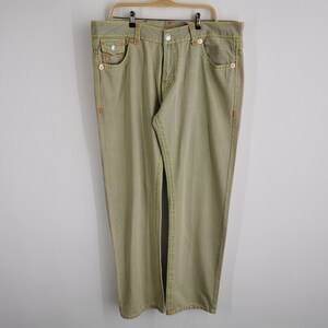 May include: Khaki-colored denim jeans with orange stitching and yellow accents. The jeans have a classic five-pocket design and are hanging on a wooden hanger. The stitching is visible on the pockets, seams, and belt loops.