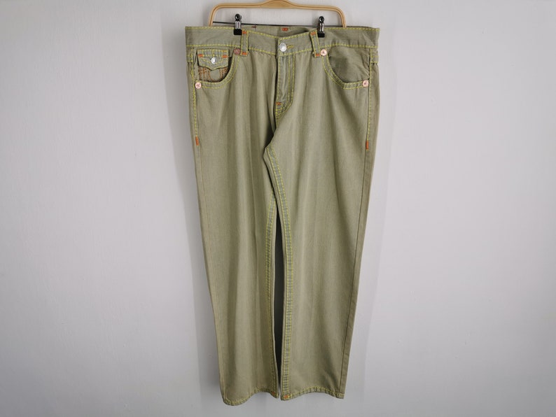 May include: Khaki-colored denim jeans with orange stitching and yellow accents. The jeans have a classic five-pocket design and are hanging on a wooden hanger. The stitching is visible on the pockets, seams, and belt loops.