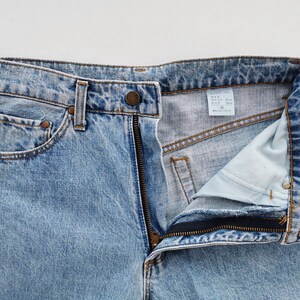 May include: Light blue denim jeans with a partially unzipped fly. The jeans have a classic five-pocket design, belt loops, and a button closure. The fabric shows a faded wash effect, and the size tag is visible.