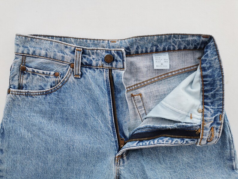 May include: Light blue denim jeans with a partially unzipped fly. The jeans have a classic five-pocket design, belt loops, and a button closure. The fabric shows a faded wash effect, and the size tag is visible.