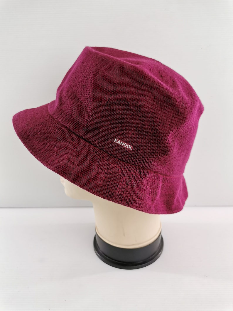 K&ouml;nnte beinhalten: Ein bordeauxfarbener Bucket Hat aus strukturiertem Stoff. Der Hut hat eine kurze Krempe und das Wort "KANGOL" in wei&szlig;en Buchstaben. Der Hut wird auf einem Schaufensterkopf pr&auml;sentiert.