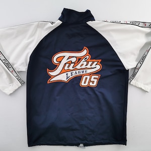 May include: A navy blue and white track jacket featuring white sleeves with a repeating FUBU logo tape down the arms. The back displays a large embroidered FUBU LEAGUE 05 graphic in orange, white, and navy blue with a high collar and drawstring hem.
