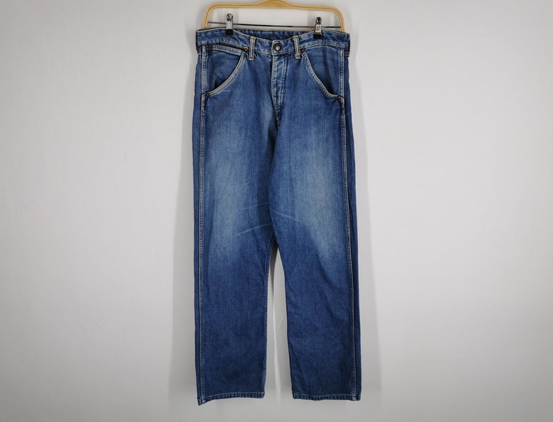 May include: Blue denim jeans hanging on a wooden hanger. The jeans have a classic five-pocket design, a button and zipper closure, and a straight leg cut. The denim has a faded wash, with lighter areas on the thighs and knees.