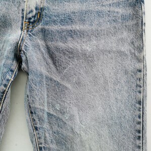 Op de afbeelding: Close-up van lichtblauwe denim jeans. De jeans heeft een vervaagde, versleten uitstraling met subtiele beschadigingen en lichtgekleurde vlekken. De stiksels zijn zichtbaar in geel en de algehele look is casual.