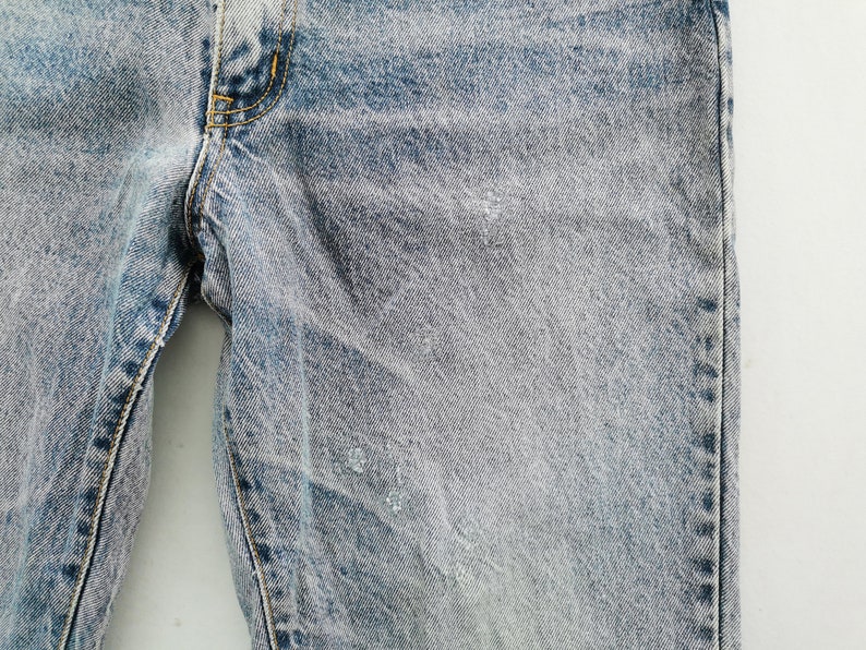 Op de afbeelding: Close-up van lichtblauwe denim jeans. De jeans heeft een vervaagde, versleten uitstraling met subtiele beschadigingen en lichtgekleurde vlekken. De stiksels zijn zichtbaar in geel en de algehele look is casual.