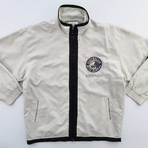 May include: A light beige zip-up jacket with black accents on the sleeves and along the zipper. The jacket features a round emblem with text that reads "Australian Sports Club Simpson."