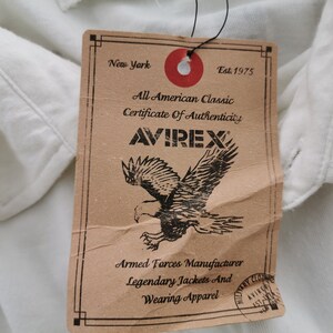 Puede incluir: Una prenda blanca con una etiqueta marr&oacute;n. La etiqueta muestra la marca "AVIREX", un gr&aacute;fico de &aacute;guila y texto como "All American Classic" y "Armed Forces Manufacturer". La etiqueta es de Nueva York.