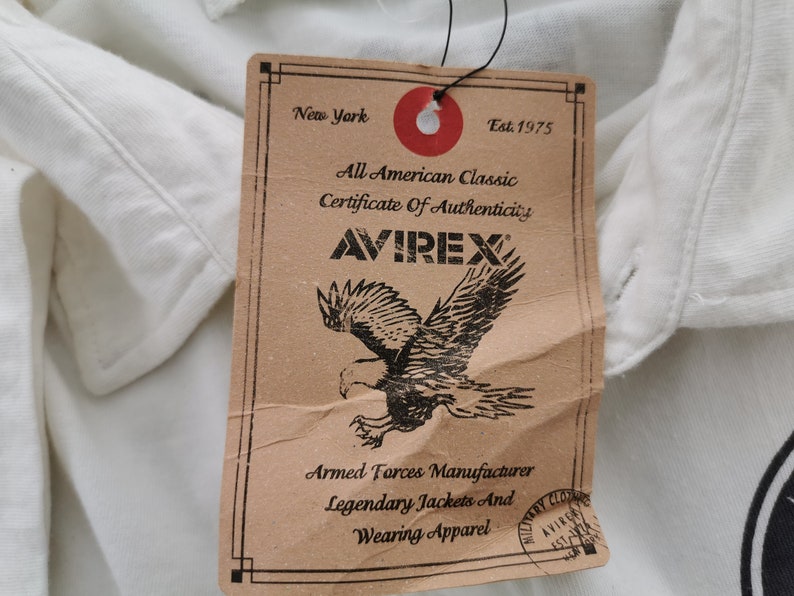 Puede incluir: Una prenda blanca con una etiqueta marr&oacute;n. La etiqueta muestra la marca "AVIREX", un gr&aacute;fico de &aacute;guila y texto como "All American Classic" y "Armed Forces Manufacturer". La etiqueta es de Nueva York.
