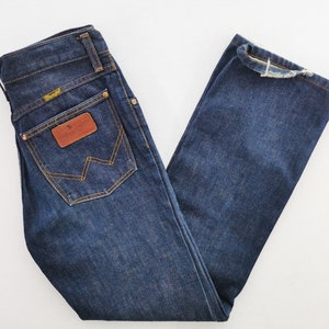 Op de afbeelding: Donkerblauwe denim jeans met een klassiek vijf-zakken ontwerp. Voorzien van een bruine leren patch en het Wrangler-logo op de achterzak. De jeans heeft een rechte pijp en is netjes opgevouwen.