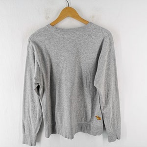 K&ouml;nnte beinhalten: Ein meliertes graues Langarm-Sweatshirt h&auml;ngt an einem Holzb&uuml;gel. Das Sweatshirt hat einen Rundhalsausschnitt und ein kleines, farbenfrohes gesticktes Design auf der unteren linken Seite. Der Hintergrund ist eine schlichte wei&szlig;e Wand.