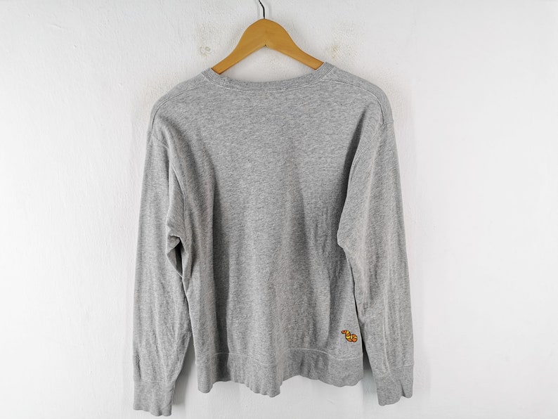 K&ouml;nnte beinhalten: Ein meliertes graues Langarm-Sweatshirt h&auml;ngt an einem Holzb&uuml;gel. Das Sweatshirt hat einen Rundhalsausschnitt und ein kleines, farbenfrohes gesticktes Design auf der unteren linken Seite. Der Hintergrund ist eine schlichte wei&szlig;e Wand.