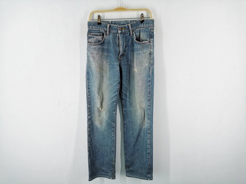 May include: Blue denim jeans with a distressed look, featuring a faded wash and a ripped knee. The jeans are hanging on a gold-colored hanger against a white background. The jeans have classic five-pocket styling.
