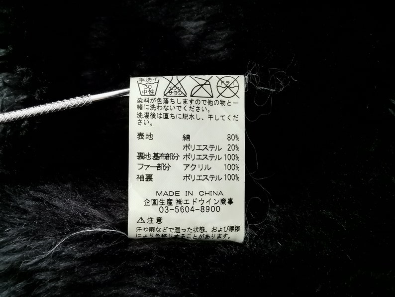 Puede incluir: Primer plano de una tela negra con una etiqueta adjunta. La etiqueta contiene texto japon&eacute;s y s&iacute;mbolos de cuidado, junto con detalles de la composici&oacute;n de la tela: 80% algod&oacute;n, 20% poli&eacute;ster y 100% acr&iacute;lico para la parte de la piel. Hecho en China.