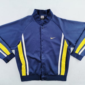 May include: A navy blue button-up jacket with yellow and white stripes on the sleeves. The jacket features a small gold Nike logo on the chest. The collar and cuffs are navy blue.