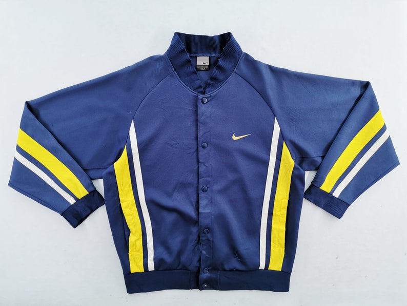 May include: A navy blue button-up jacket with yellow and white stripes on the sleeves. The jacket features a small gold Nike logo on the chest. The collar and cuffs are navy blue.
