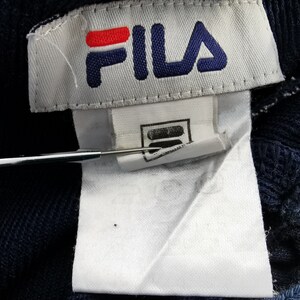 Puede incluir: Primer plano de una etiqueta de tela azul marino con el logotipo de FILA en rojo y azul. La etiqueta est&aacute; adherida a una tela azul marino. Una herramienta plateada apunta a la etiqueta.