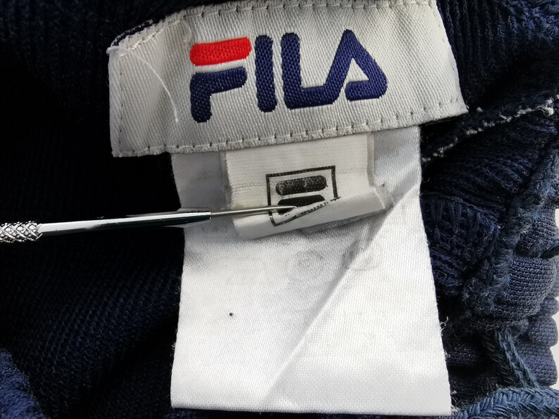 Puede incluir: Primer plano de una etiqueta de tela azul marino con el logotipo de FILA en rojo y azul. La etiqueta est&aacute; adherida a una tela azul marino. Una herramienta plateada apunta a la etiqueta.