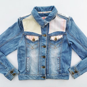 May include: A light-wash denim jacket with patterned fabric patches on the shoulders and pockets. The jacket has a button-up front, two chest pockets, and long sleeves with buttoned cuffs. The patches feature floral and gingham patterns.