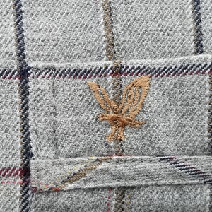 以下が含まれることがあります： 格子柄のグレーのツイード生地のクローズアップ。茶色の刺繍されたワシのエンブレムで飾られた小さなポケットが見えます。生地は、濃い青、赤、茶色のアクセントラインでテクスチャ加工されています。