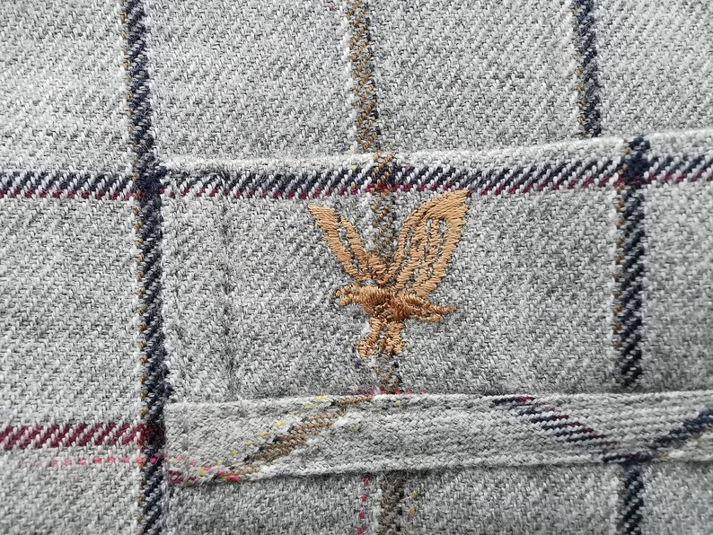 以下が含まれることがあります： 格子柄のグレーのツイード生地のクローズアップ。茶色の刺繍されたワシのエンブレムで飾られた小さなポケットが見えます。生地は、濃い青、赤、茶色のアクセントラインでテクスチャ加工されています。