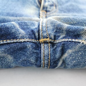 May include: Close-up of blue denim jeans, showing the fly, seams, and distressed details. The stitching is a contrasting tan color. The denim has a faded, worn appearance.