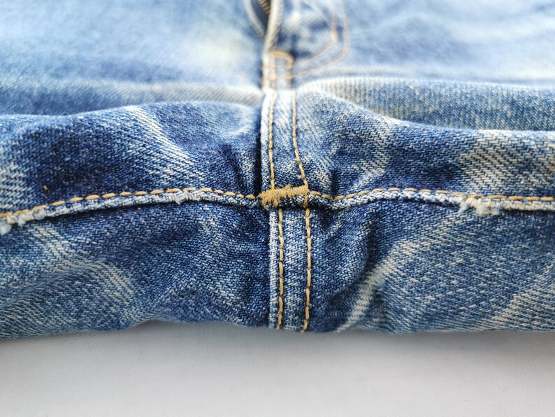 May include: Close-up of blue denim jeans, showing the fly, seams, and distressed details. The stitching is a contrasting tan color. The denim has a faded, worn appearance.