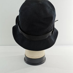 May include: A black cloche hat with a wide brim and a black ribbon band. The hat is made of a woven material and is displayed on a mannequin head. The hat has a classic, vintage style.