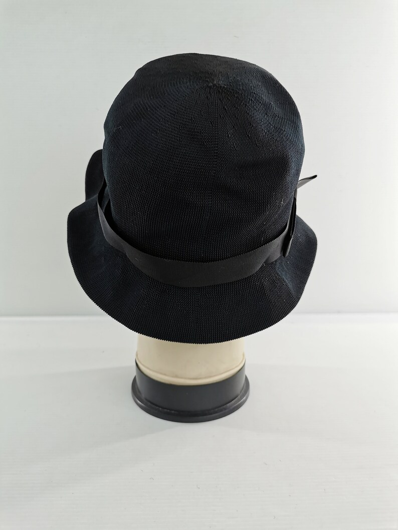 May include: A black cloche hat with a wide brim and a black ribbon band. The hat is made of a woven material and is displayed on a mannequin head. The hat has a classic, vintage style.