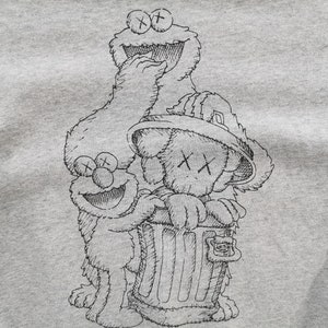 K&ouml;nnte beinhalten: Ein hellgraues Sweatshirt mit einer schwarz-wei&szlig;en Strichzeichnung der Sesamstra&szlig;en-Charaktere Kr&uuml;melmonster und Elmo sowie einer Figur im KAWS-Stil. Die Figuren sind gruppiert, wobei die KAWS-Figur einen M&uuml;lleimer h&auml;lt.