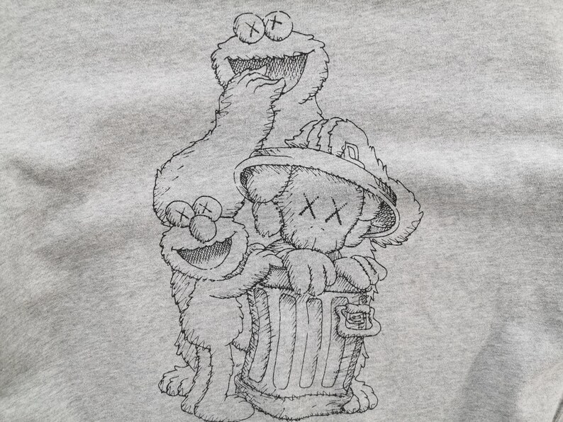 K&ouml;nnte beinhalten: Ein hellgraues Sweatshirt mit einer schwarz-wei&szlig;en Strichzeichnung der Sesamstra&szlig;en-Charaktere Kr&uuml;melmonster und Elmo sowie einer Figur im KAWS-Stil. Die Figuren sind gruppiert, wobei die KAWS-Figur einen M&uuml;lleimer h&auml;lt.