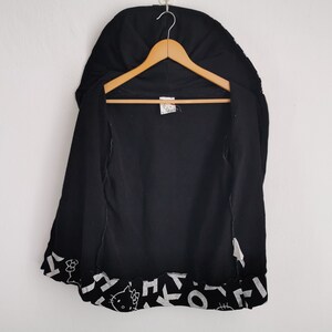 May include: Black zip-up hoodie with a hood, hanging on a wooden hanger. The bottom hem features a repeating pattern of white shapes and a Hello Kitty graphic. The hoodie is open, revealing the interior.