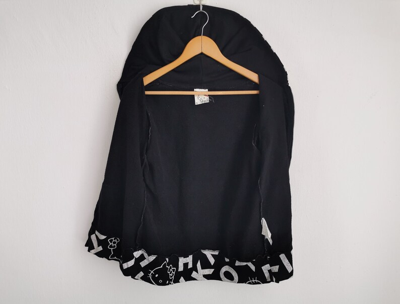 May include: Black zip-up hoodie with a hood, hanging on a wooden hanger. The bottom hem features a repeating pattern of white shapes and a Hello Kitty graphic. The hoodie is open, revealing the interior.