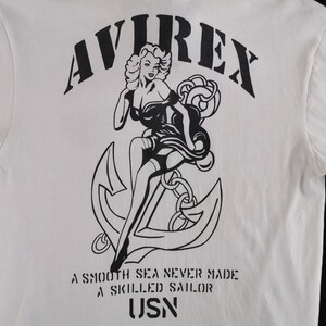 Puede incluir: Camiseta blanca con un gr&aacute;fico negro de una mujer, un ancla y el texto "AVIREX". Debajo del gr&aacute;fico, el texto dice "A SMOOTH SEA NEVER MADE A SKILLED SAILOR" y "USN".