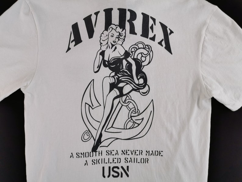 Puede incluir: Camiseta blanca con un gr&aacute;fico negro de una mujer, un ancla y el texto "AVIREX". Debajo del gr&aacute;fico, el texto dice "A SMOOTH SEA NEVER MADE A SKILLED SAILOR" y "USN".