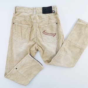 May include: Beige corduroy pants featuring a high-waisted design with two back pockets, a black rectangular patch on the waistband, and a red embroidered graphic on the right back pocket. The pants include metal rivets and a button closure.