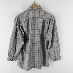 以下が含まれることがあります： 黒と茶色のチェック柄のグレーの長袖ボタンダウンシャツ。 シャツは白い背景に木製のハンガーに吊るされています。 袖口には2つのボタンが付いています。