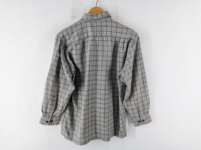 以下が含まれることがあります： 黒と茶色のチェック柄のグレーの長袖ボタンダウンシャツ。 シャツは白い背景に木製のハンガーに吊るされています。 袖口には2つのボタンが付いています。