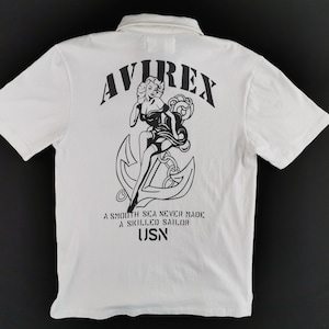 Puede incluir: Polo blanco con un gr&aacute;fico negro en la espalda que presenta a una mujer, un ancla y el texto "AVIREX". La camisa tambi&eacute;n tiene el texto "A SMOOTH SEA NEVER MADE A SKILLED SAILOR" y "USN".