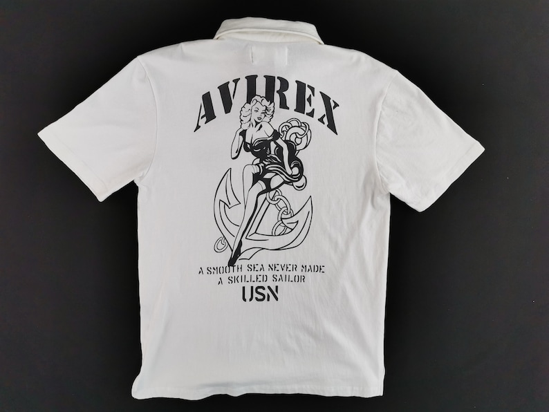 Puede incluir: Polo blanco con un gr&aacute;fico negro en la espalda que presenta a una mujer, un ancla y el texto "AVIREX". La camisa tambi&eacute;n tiene el texto "A SMOOTH SEA NEVER MADE A SKILLED SAILOR" y "USN".
