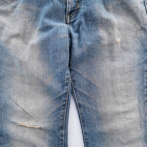 May include: Faded blue denim jeans with a distressed look. The jeans have a worn appearance with a ripped knee and faded patches. The classic five-pocket design is visible.